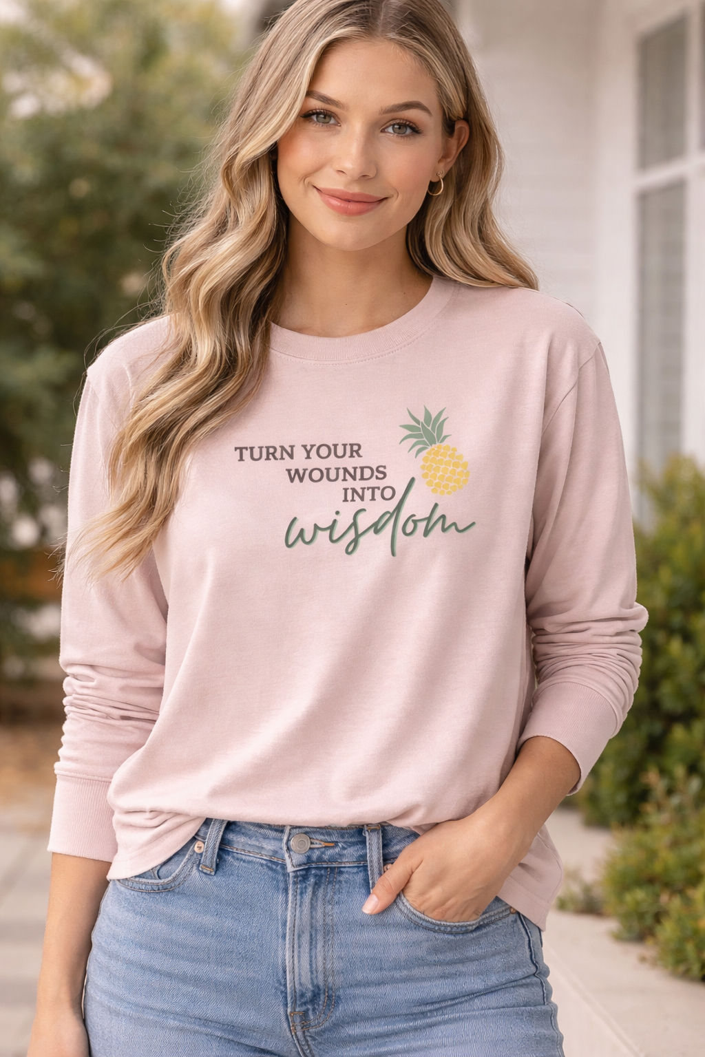 Woman model wearing blush pink long sleeve shirt with “Turn Your Wounds Into Wisdom” pineapple graphic, styled with jeans for Hopeful Hearts Clothing Co website
