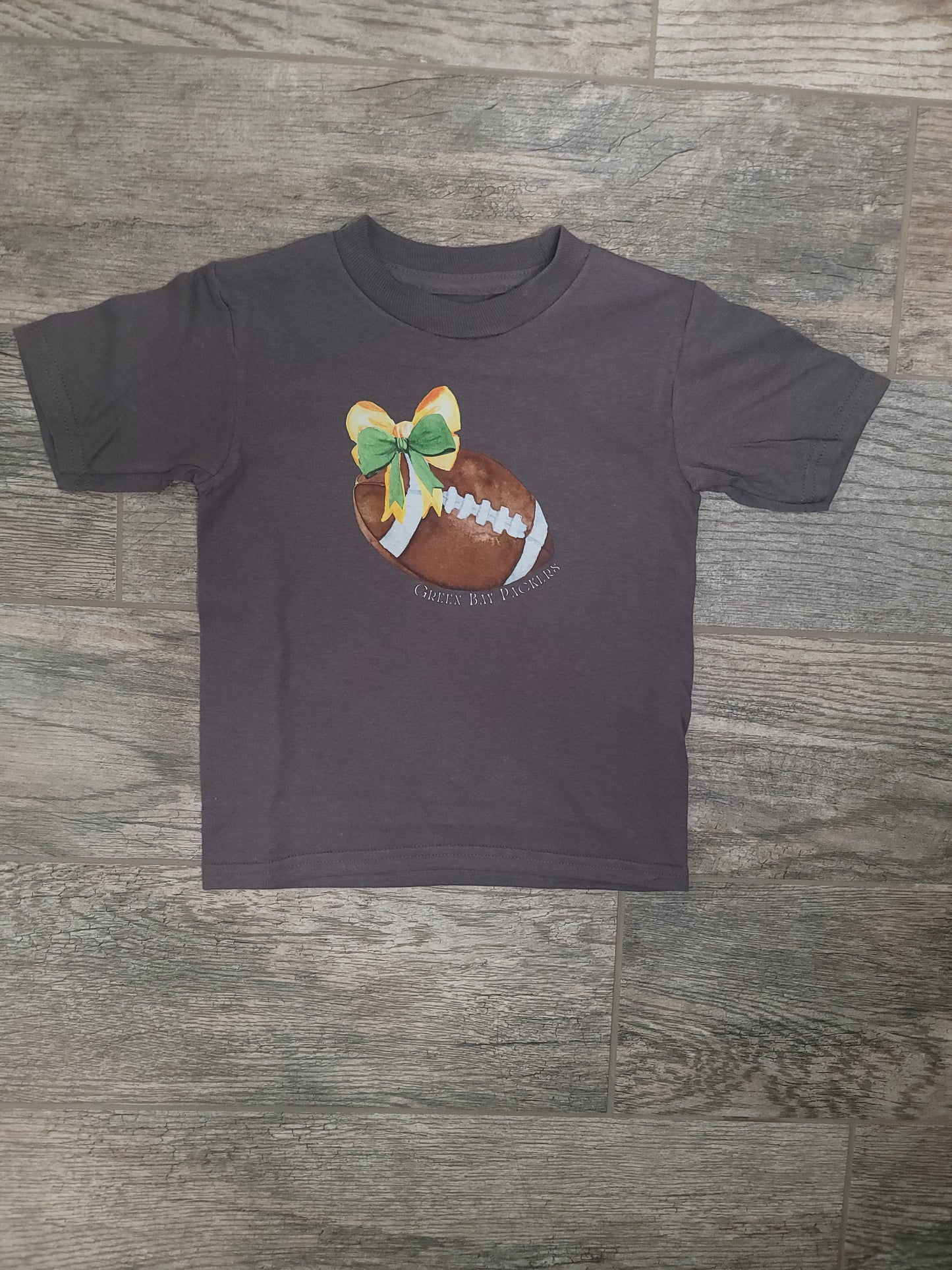 Flat lay of Green Bay Packers Toddler Tee styled with bow and football, showing team pride outfit option