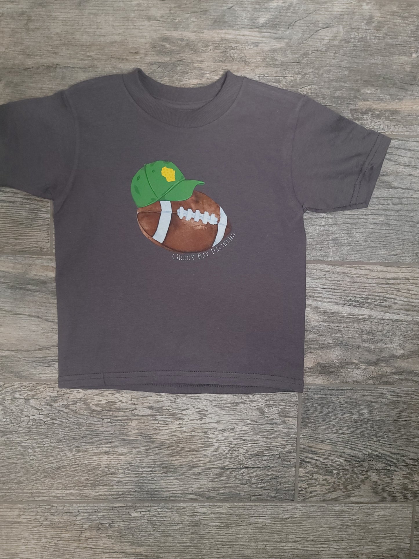 Flat lay of Green Bay Packers Toddler Tee styled with baseball cap and football, showcasing team pride outfit option