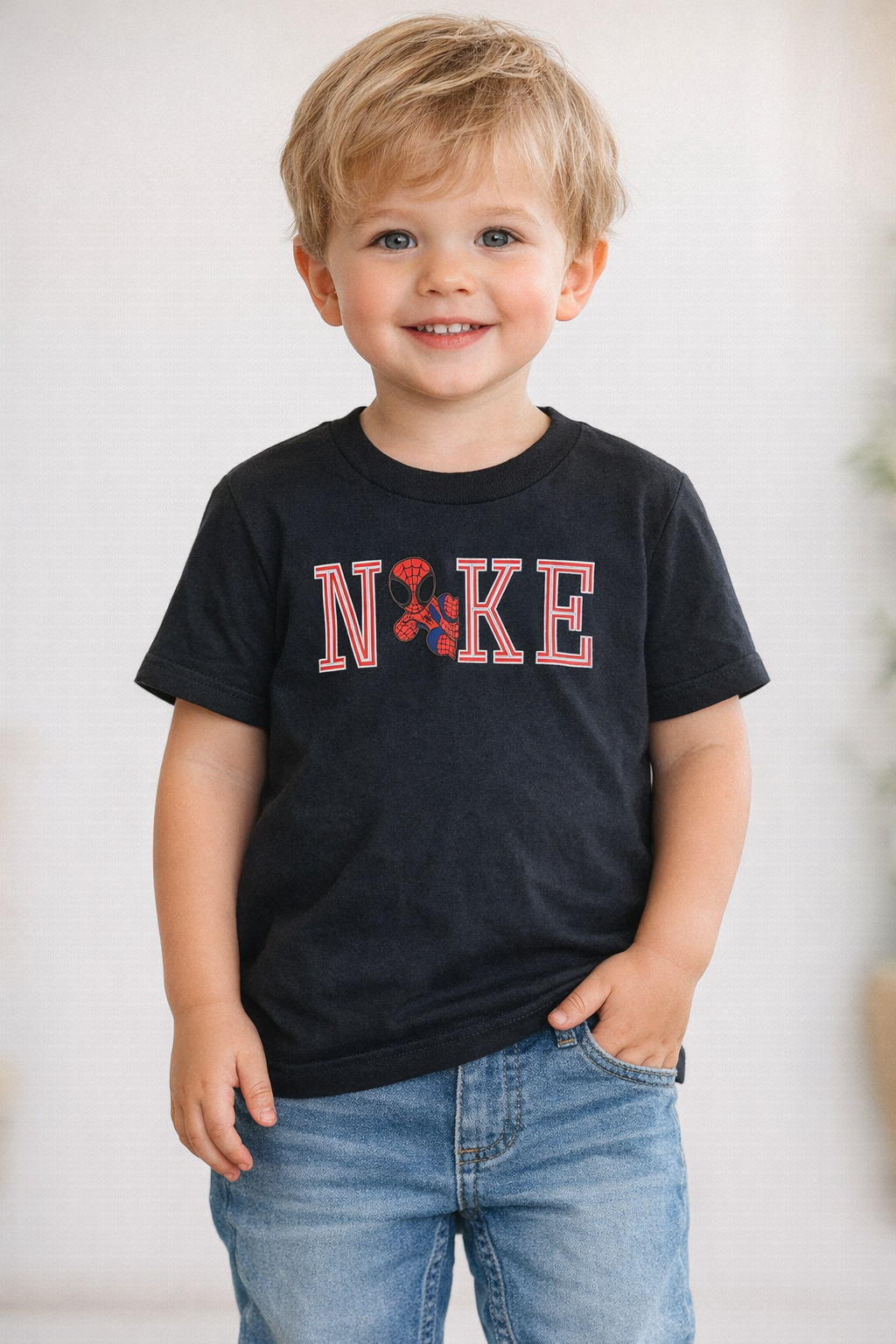 Toddler wearing a black graphic t-shirt with red and white lettering and a spider-inspired design, standing in a neutral studio setting.