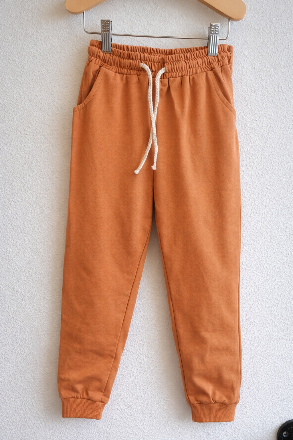 Burnt orange kids jogger pants displayed on a hanger against a white wall, featuring an elastic waistband with drawstring and ribbed ankle cuffs