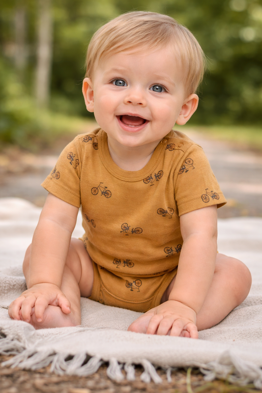 6–12 month baby modeling mustard bicycle print onesie outdoors.