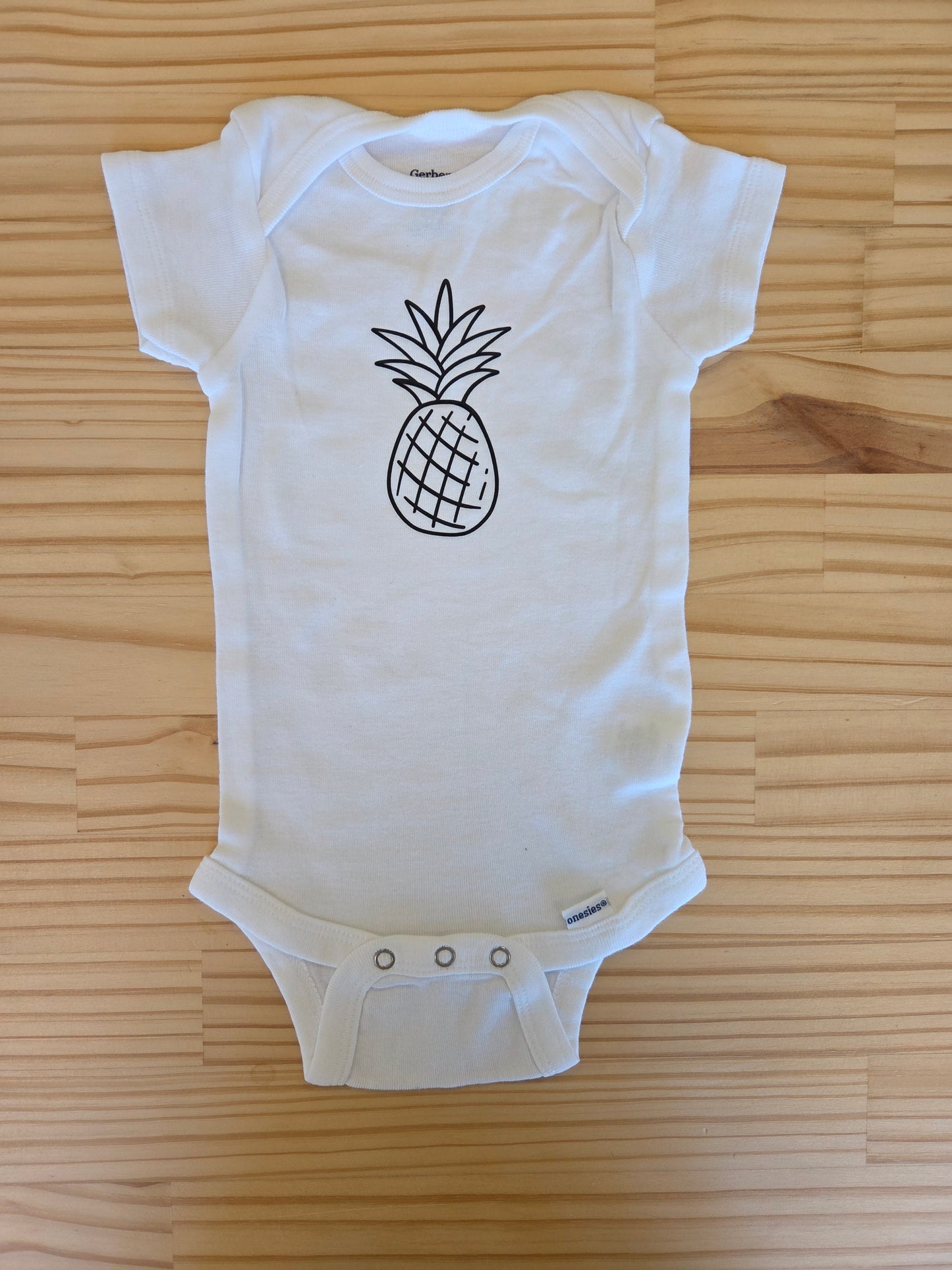Flat lay of white baby onesie size 3–6 months with black pineapple print, symbolizing infertility awareness, rainbow babies, and hope