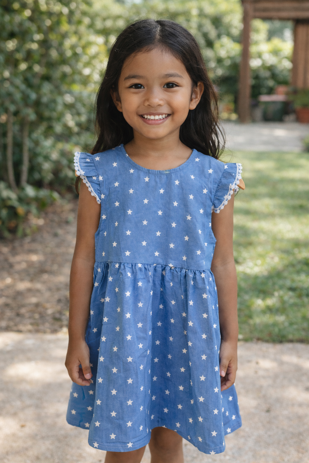 Six-year-old girl standing outdoors wearing a smooth blue chambray dress with white star print and flutter sleeves.
