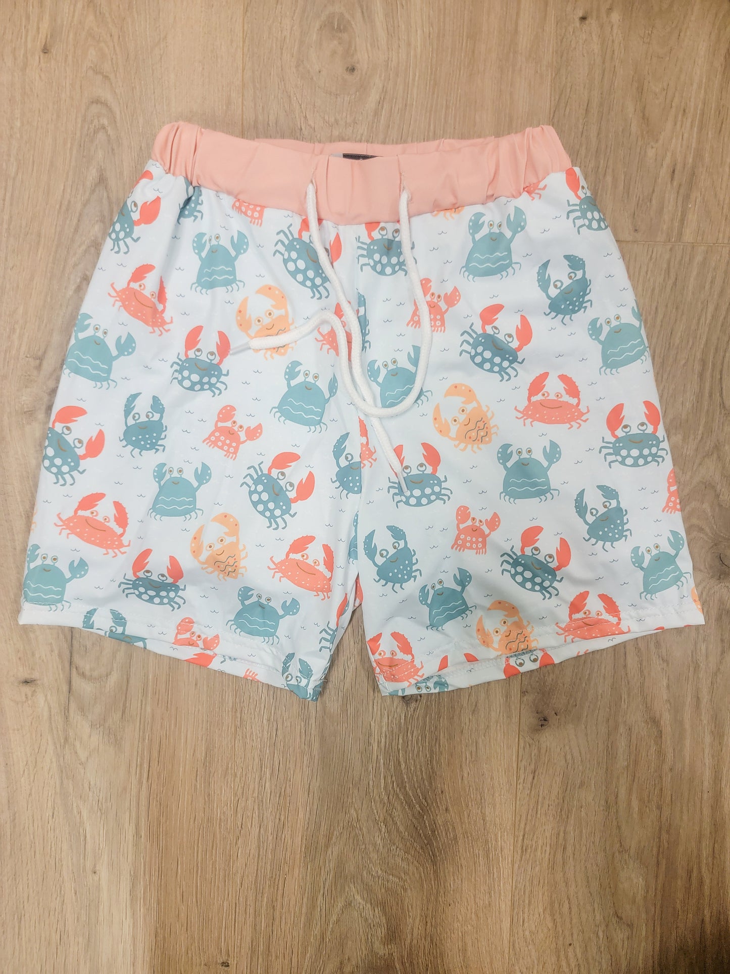 Flat lay of boys crab print swim trunks featuring coral, teal, and blue crabs with a soft pink waistband