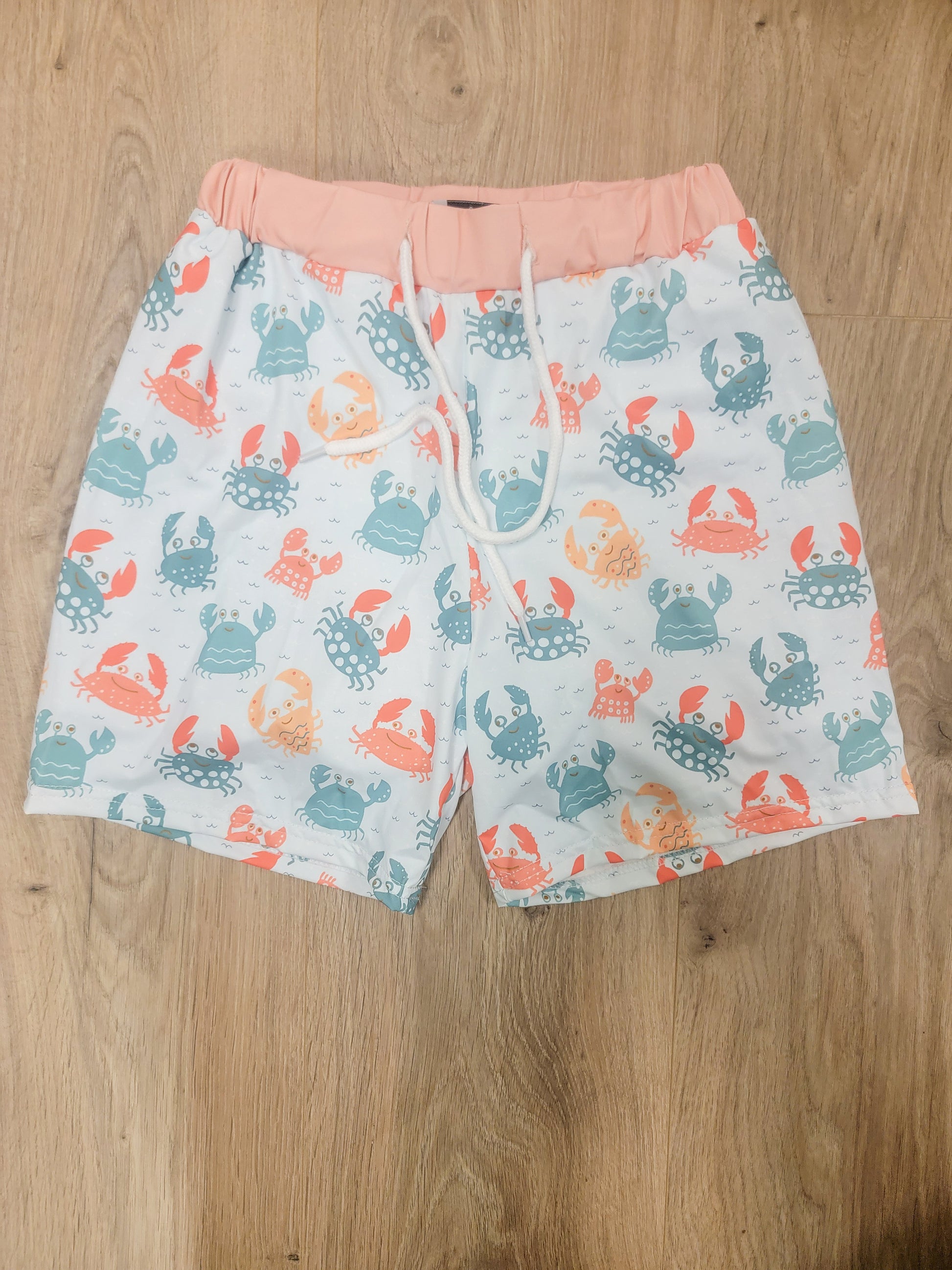 Flat lay of boys crab print swim trunks featuring coral, teal, and blue crabs with a soft pink waistband