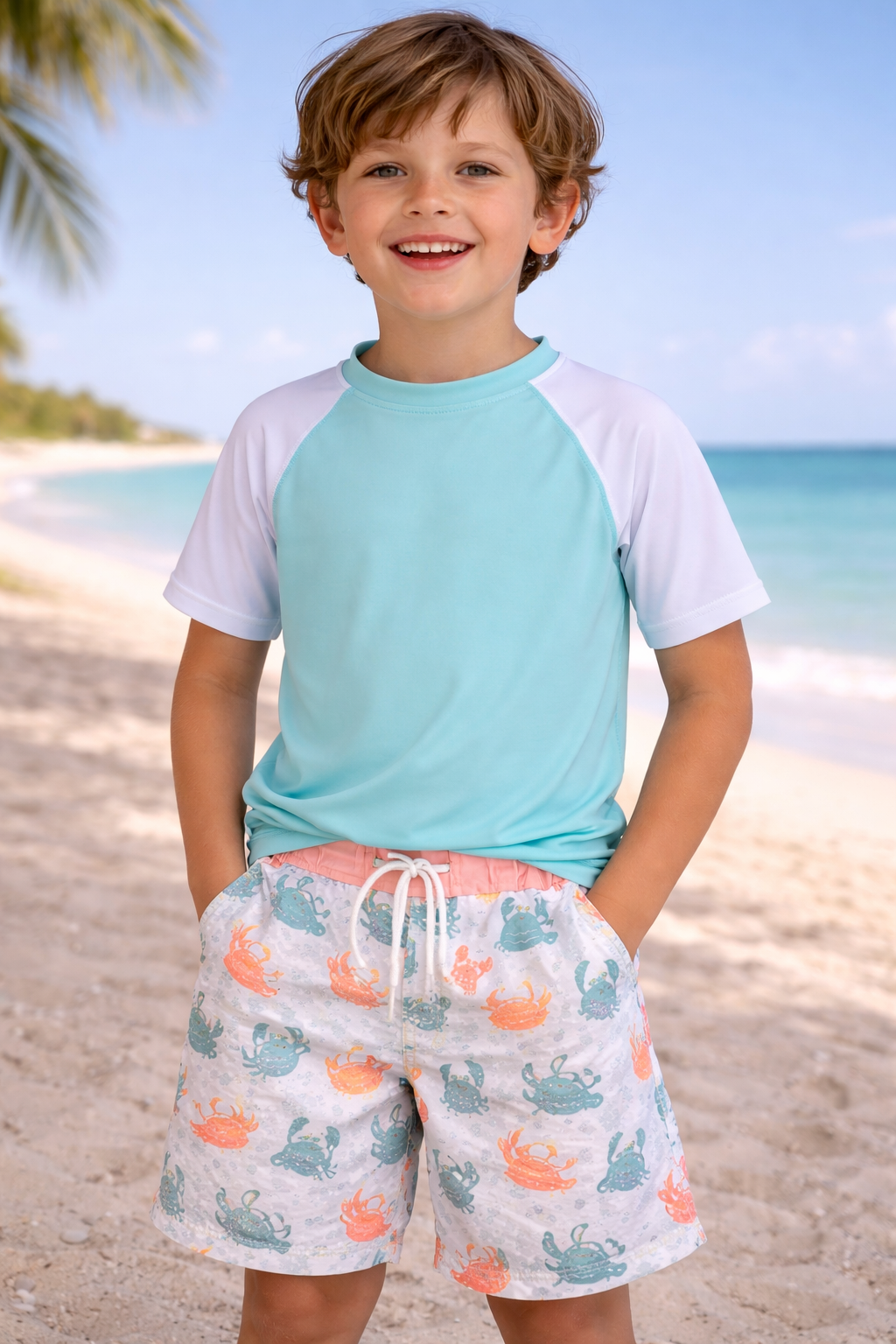 Young child wearing crab print swim trunks with a solid aqua swim top, shown on a beach for kids summer swimwear styling
