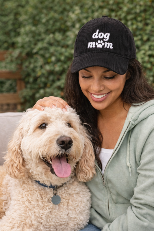 Woman wearing black corduroy “dog mom” hat while sitting with her dog in an outdoor lifestyle setting