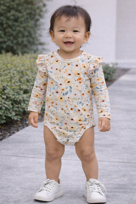 Toddler under 18 months wearing a cream floral long sleeve onesie with ruffle shoulders, styled on a standing model for baby boutique clothing