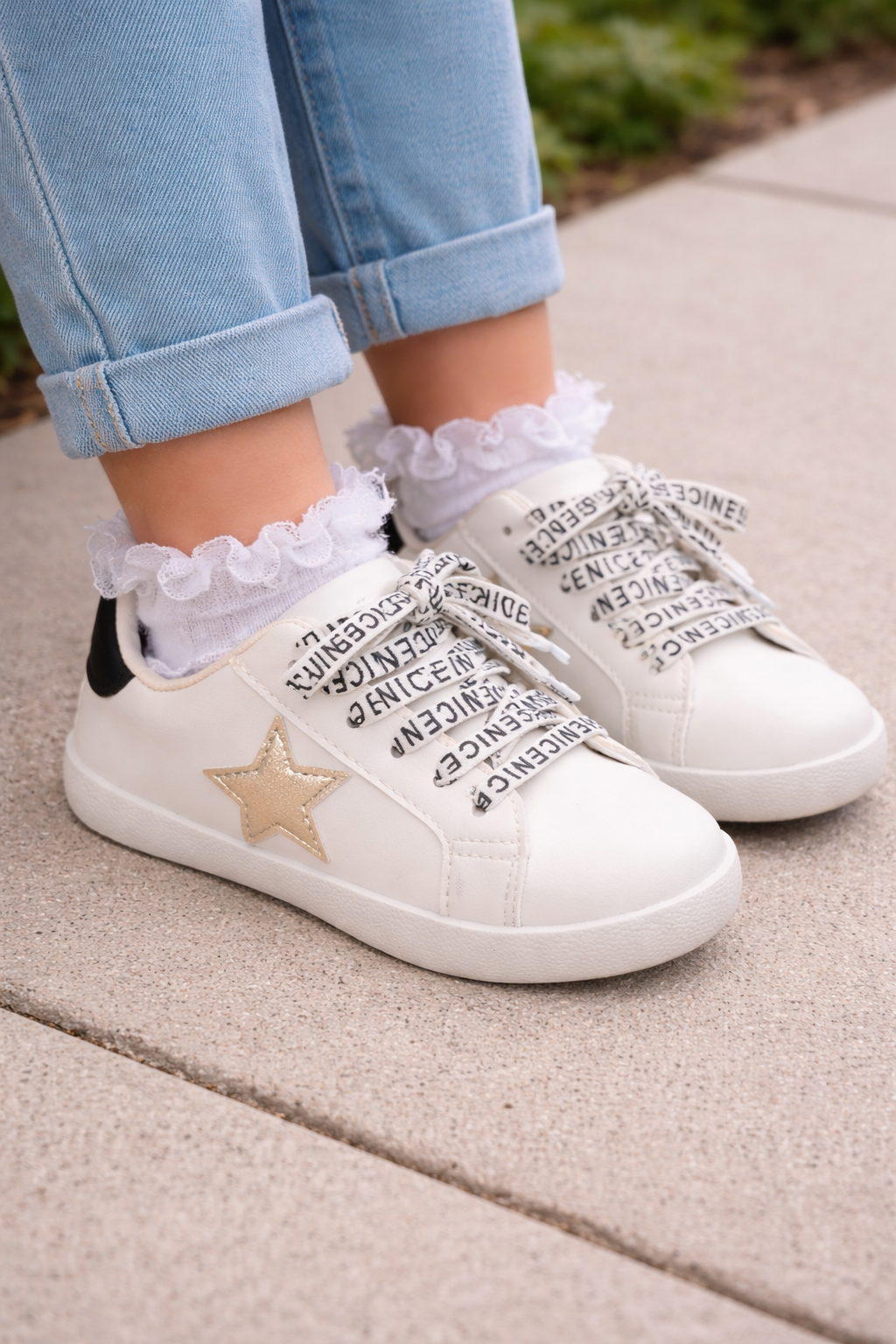 Young girl wearing white youth sneakers with gold star detail and black heel accent, styled with cuffed jeans and lace socks, shoes shown close-up as the focal point