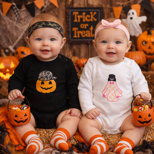 Six-month-old baby boy and girl models wearing Halloween long sleeve bodysuits, featuring a pumpkin design on black and a pink ghost design on white, styled with a festive Halloween background