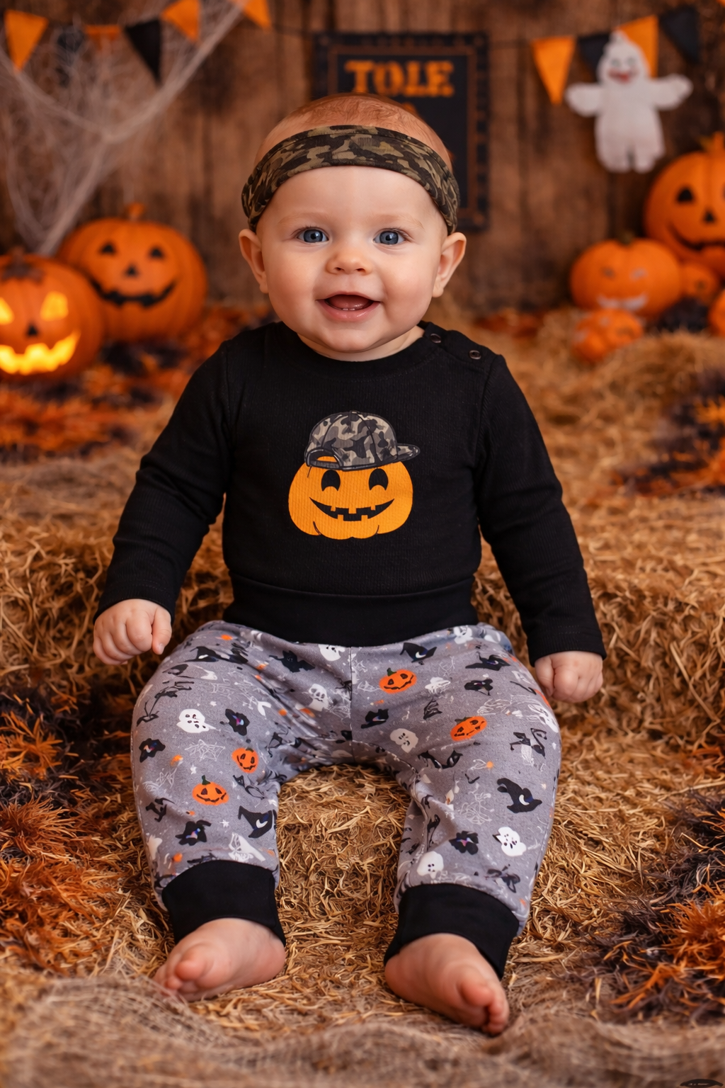 Baby boy model wearing a black long sleeve pumpkin Halloween bodysuit paired with gray spooky print pants, styled in a festive Halloween fall background.