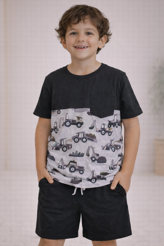 Older child wearing a black and gray colorblock t-shirt with construction vehicle print and matching black drawstring shorts, standing in a neutral studio setting.