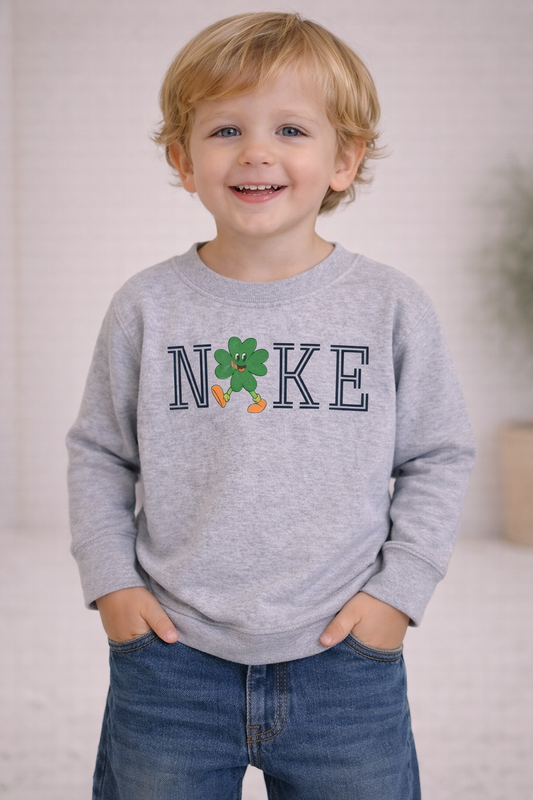 Toddler wearing a heather gray sweatshirt featuring a shamrock graphic and “NKE” text, standing in a neutral studio setting.