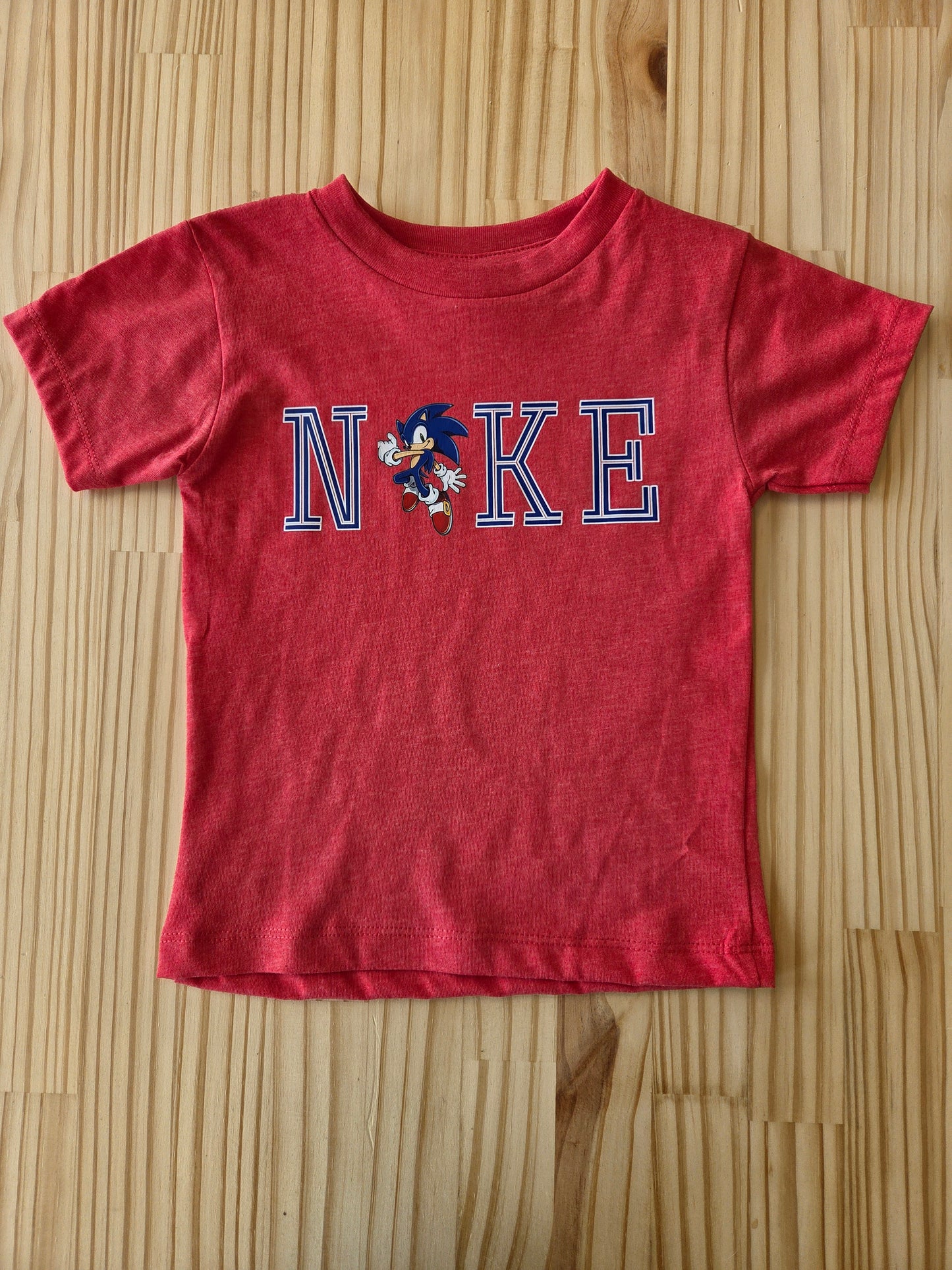 Red toddler short sleeve t-shirt laid flat, featuring “NKE” text and a cartoon hedgehog graphic on the front.