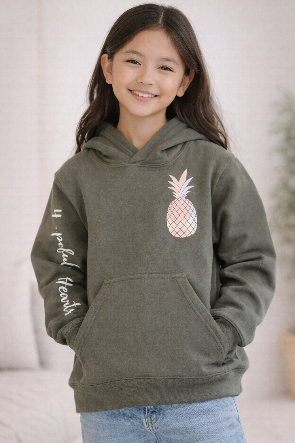 Youth wearing an olive green hoodie with pineapple graphic and “Hopeful Hearts” text on the sleeve, smiling in a neutral studio setting.