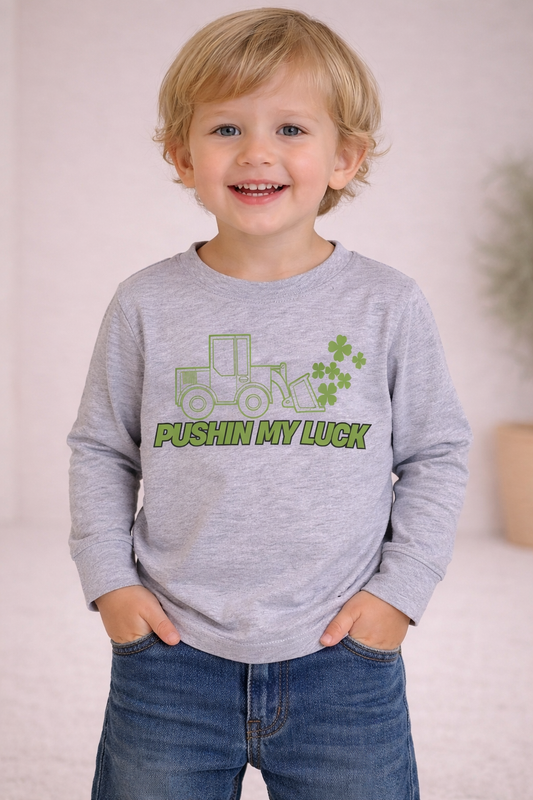 Toddler wearing a heather gray long sleeve shirt with green tractor graphic and “Pushin My Luck” text, standing in a neutral studio setting.