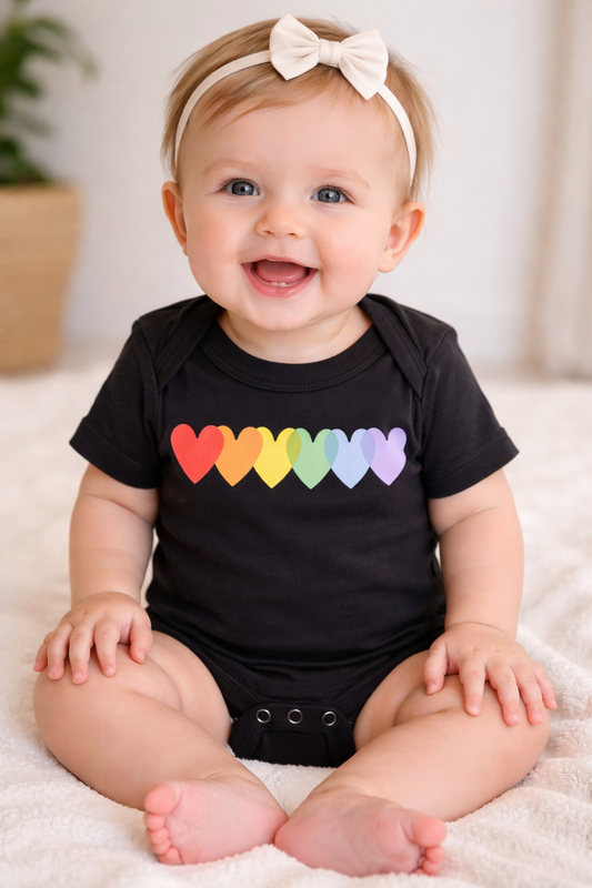 Infant wearing a black rainbow hearts onesie, sitting on a soft white blanket, smiling in a neutral studio setting.