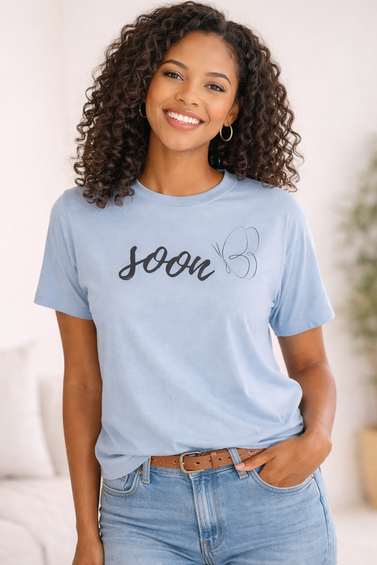 Adult woman wearing a light blue graphic t-shirt with “soon” script and butterfly design, smiling in a neutral studio setting.