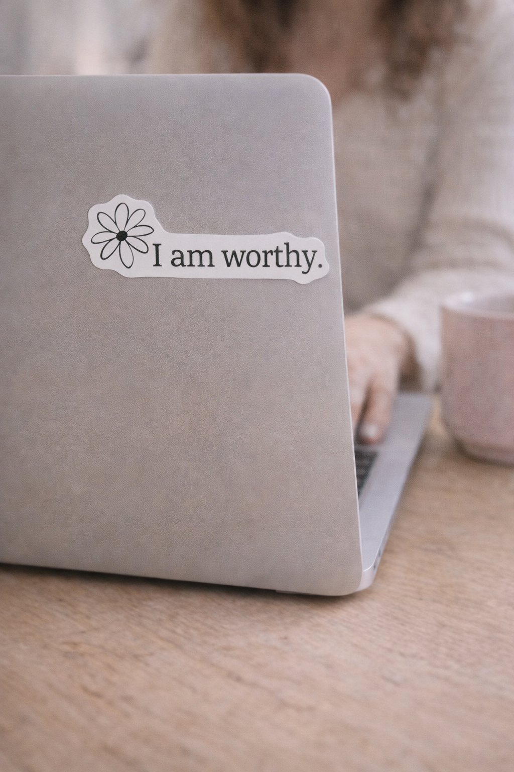 “I am worthy” vinyl sticker with daisy design placed on the cover of a silver laptop, shown in a cozy workspace setting with a coffee mug, illustrating real-life use and scale
