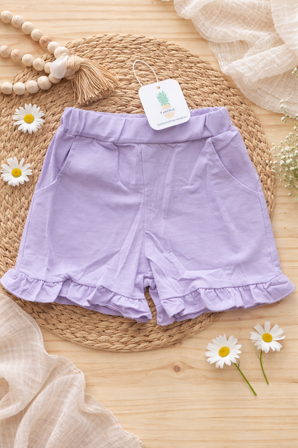 Lavender ruffled baby shorts styled in a flat lay with woven textures, highlighting elastic waistband and flutter hem