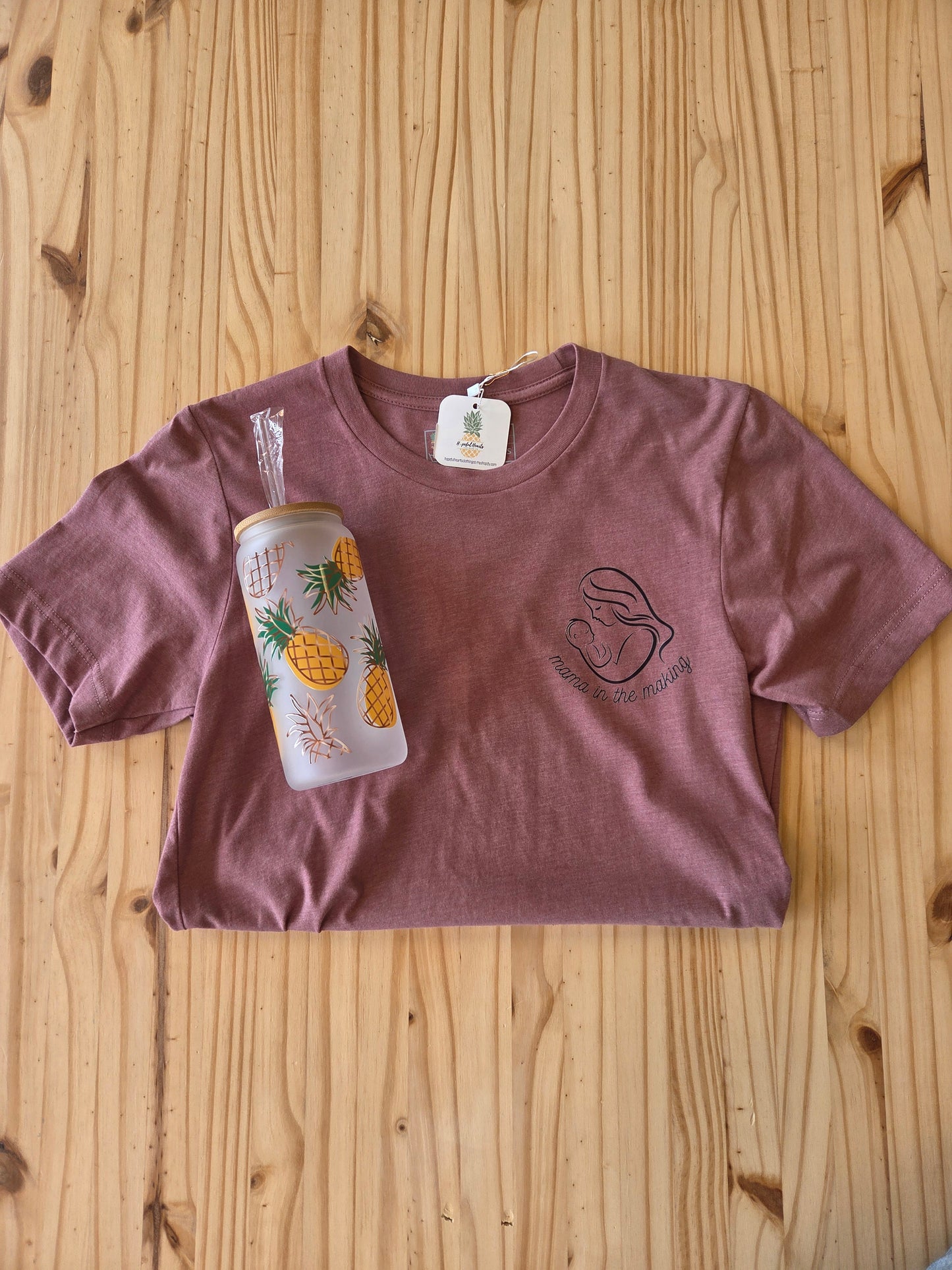 Flat lay of a “Mama in the Making” graphic t-shirt paired with a pineapple tumbler, styled as a gift bundle representing infertility support and hope, arranged on a light neutral background for a soft, meaningful aesthetic.