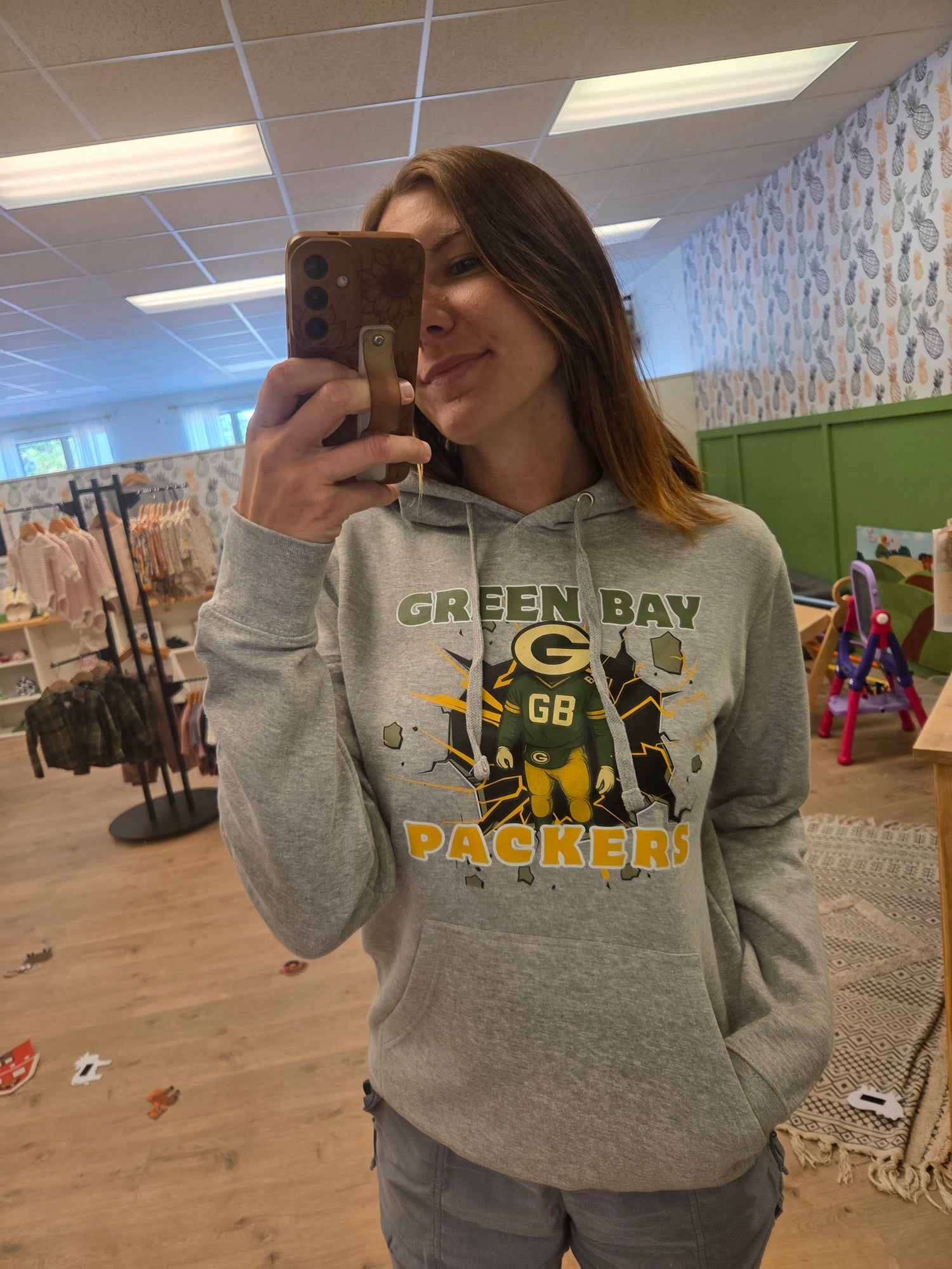 A woman taking a mirror selfie in a clothing store, wearing a light gray hoodie with a 'Green Bay Packers' graphic on the front.