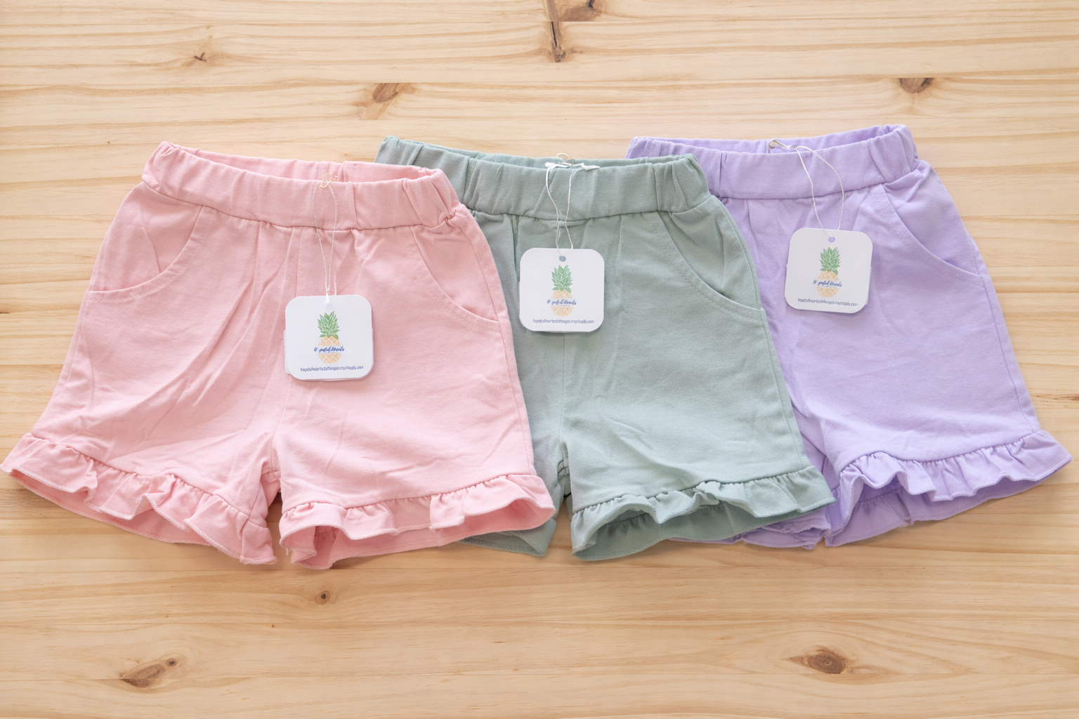 Three pairs of ruffled baby shorts displayed side by side in pink, sage green, and lavender