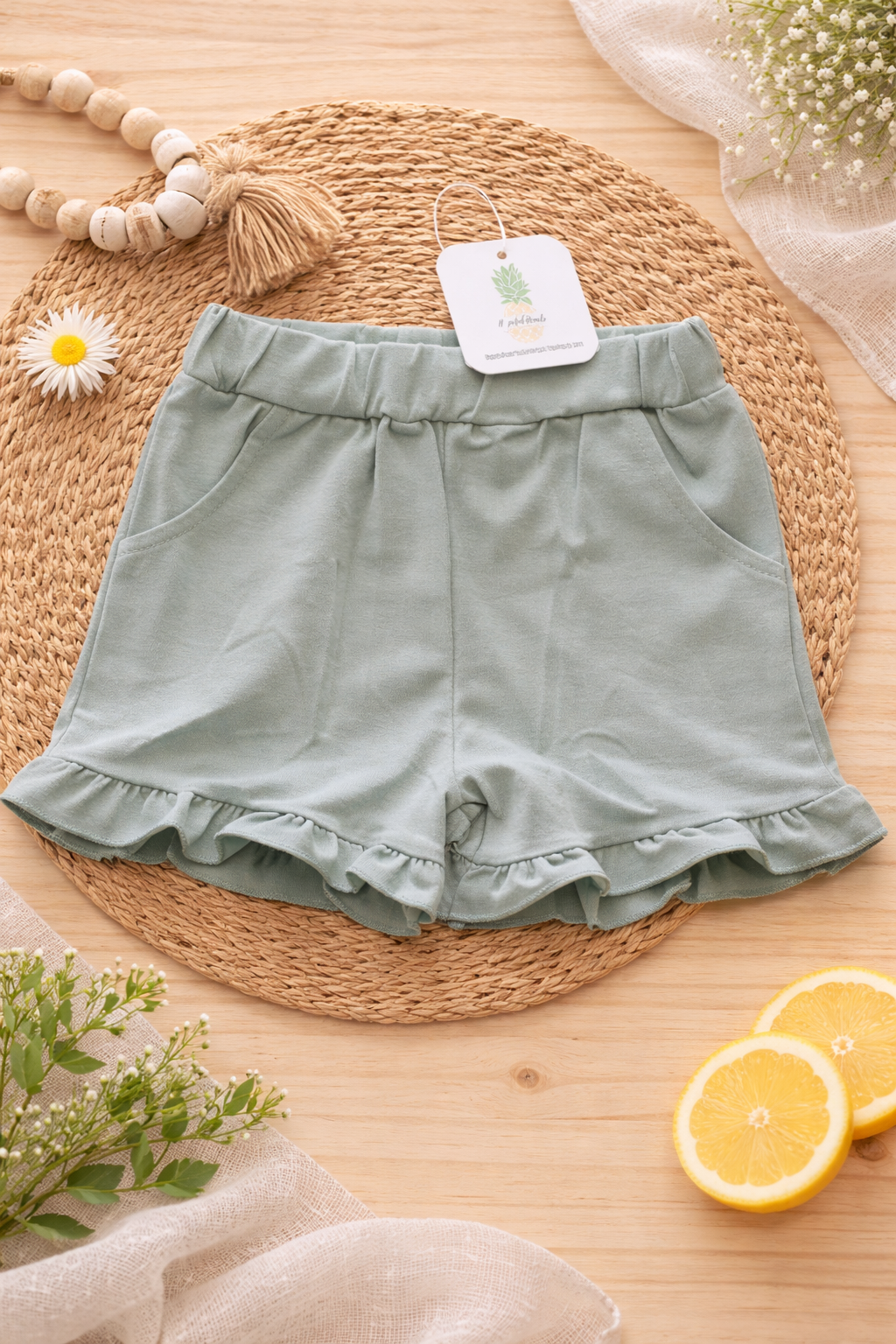Sage green ruffled baby shorts displayed in a styled flat lay, showing soft fabric, pockets, and ruffled hem detail