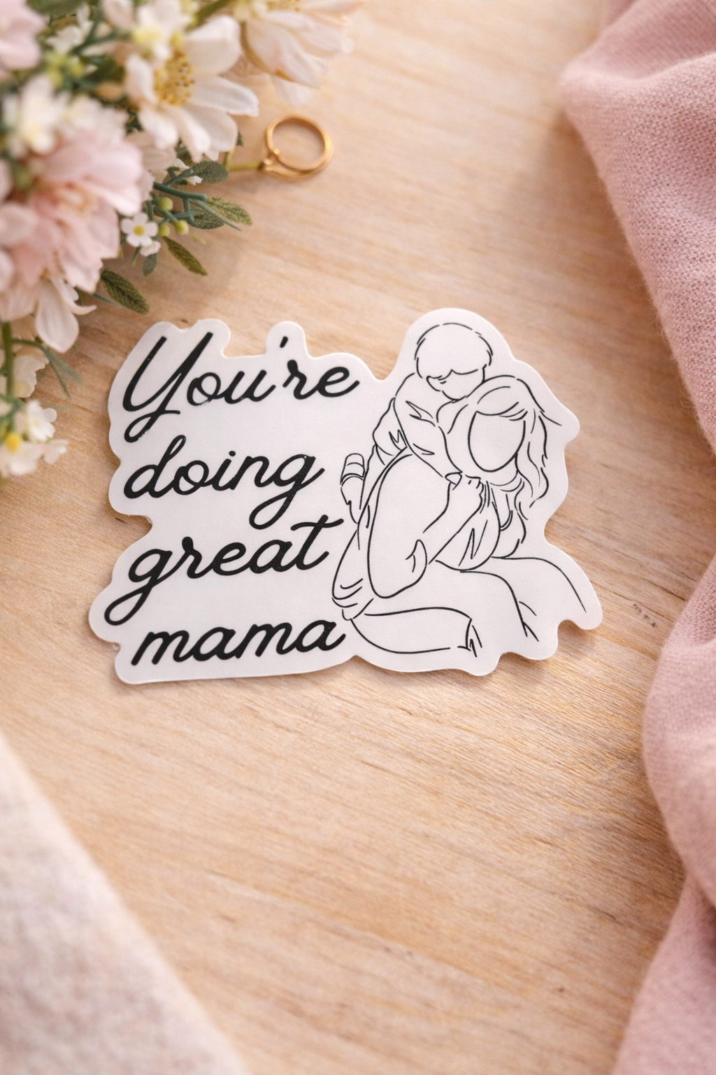 You’re Doing Great Mama vinyl sticker featuring an illustrated mother and child, styled on a light wood surface with soft florals and neutral accents.