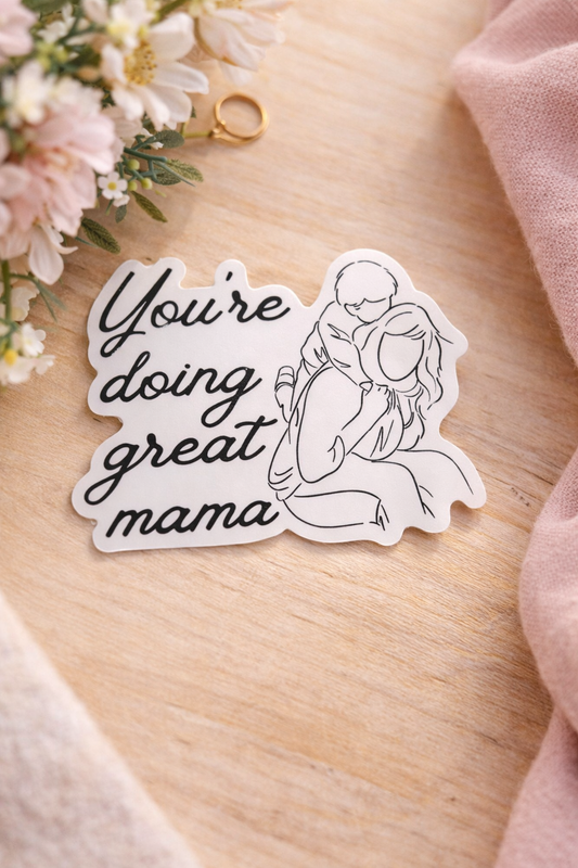 You’re Doing Great Mama vinyl sticker featuring an illustrated mother and child, styled on a light wood surface with soft florals and neutral accents.