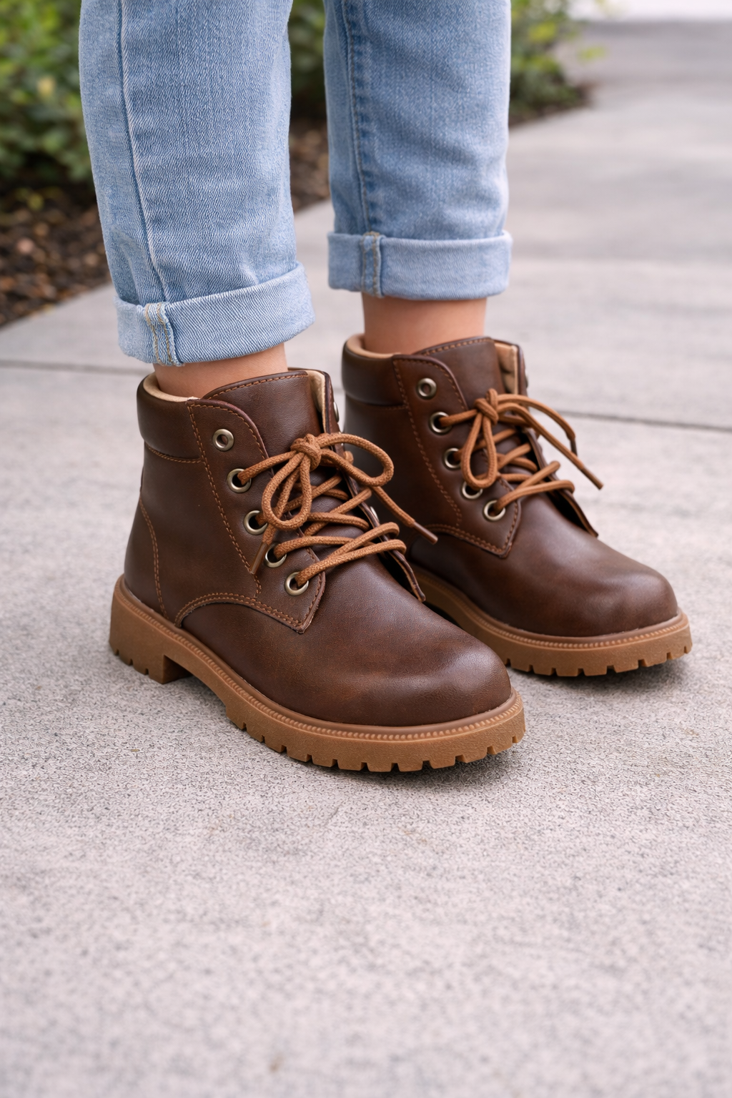 Youth model wearing brown lace-up combat boots with tan rubber soles, shown on feet with cuffed jeans to highlight fit and style